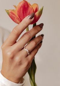 Złoty pierścionek o falistym wzorze z pięcioma błyszczącymi, przezroczystymi kamieniami. Dłoń trzymająca czerwoną i żółtą tulipan z zielonymi liśćmi. Neutralne tło.