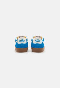 Zapatillas de gamuza azul con acentos blancos, con el logo "nike" en el talón y una suela de goma marrón. Vista trasera que muestra detalles de costura.