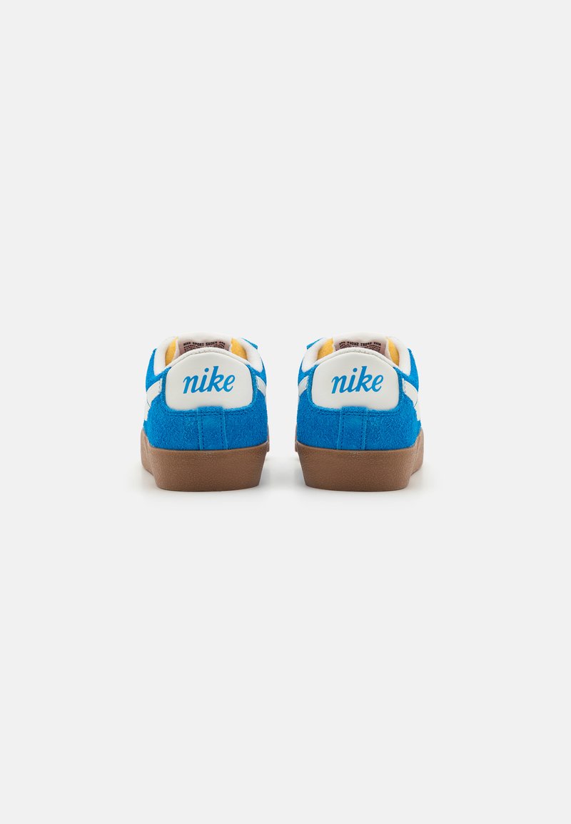 Baskets bleu suédé avec des accents blancs, comportant le logo "nike" sur le talon et une semelle extérieure en caoutchouc marron. Vue arrière montrant les détails de couture.