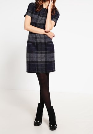 Kobieta nosząca szarą i granatową kraciastą sukienkę za kolano z krótkim rękawem, czarne rajstopy oraz czarne botki z białymi noskami.