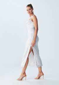 White sequin dress with a fitted design, slit on the left side, and thin straps. Worn with nude high-heel sandals.