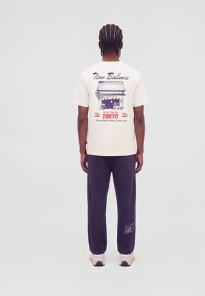 Мъж с плитки носи кремава тениска на New Balance с графика на магазина в Токио и тъмносини спортни панталони с лого, стои с гръб към зрителя.