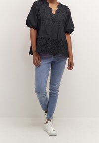 Musta brodeerattu bleiser, jossa on aaltoileva helma, lyhyet puffihihat ja syvä V-pääntie, yhdistettynä vaaleansinisiin skinny-farkkuihin ja valkoisiin lenkkitossuihin.