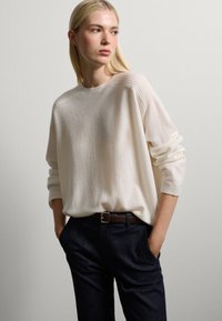 Maglione lavorato a maglia di colore crema, con scollo rotondo e dettagli a coste, abbinato a pantaloni in denim scuro e una cintura in pelle marrone.