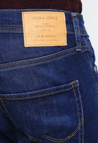 Mörkblå denimjeans med en beige läderbrandpatch, gula sömmar och en klassisk femficksdesign.