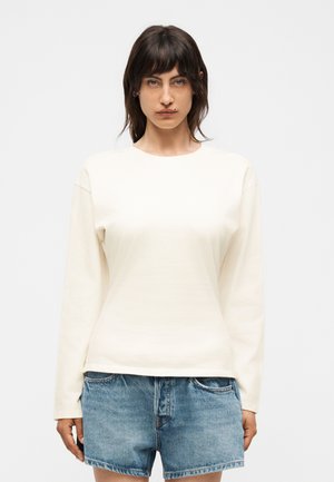 Γυναίκα με σκούρα μαλλιά που φοράει μια απλή μακρυμάνικη μπλούζα σε απαλό εκρού χρώμα και μπλε τζιν σορτς, στέκεται μπροστά σε λευκό φόντο.