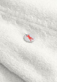 Wollene stof in witte textuur met een zilveren cirkelvormig label met een rode hondsilhouet. Minimalistisch ontwerp met een glanzende afwerking.