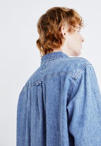Cămașă din denim de culoare albastră deschisă, cu un yoke la spate și un pliu subtil. Prezintă un guler clasic și o croială relaxată, realizată dintr-un material texturat.