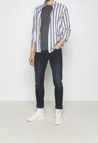 Conjunto de hombre que incluye una camisa de botones a rayas en blanco y gris, una camiseta gris oscuro, jeans oscuros y zapatillas blancas.