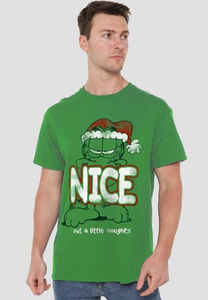Mann trägt ein grünes T-Shirt mit einer cartoonhaften Katze in einem Santa-Hut und dem Text "NICE, aber ein wenig unartig" auf der Vorderseite vor einem einfarbigen Hintergrund.