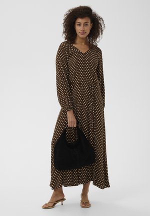 Femme aux cheveux bouclés portant une longue robe marron à pois, tenant un sac à main noir et portant des sandales à talons beige, debout sur un fond blanc.