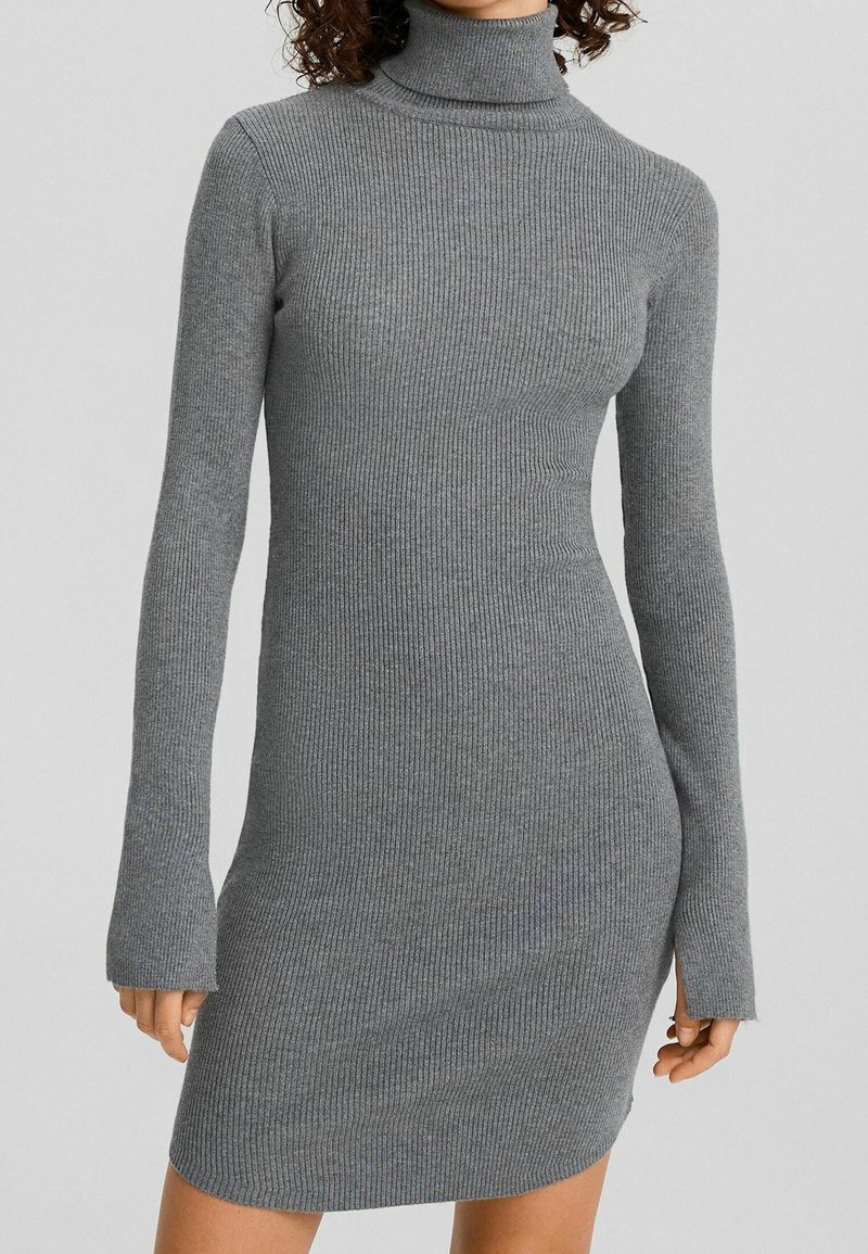 Femme portant une robe-pull ajustée en maille côtelée grise à col roulé, avec des manches longues dépassant légèrement les poignets.