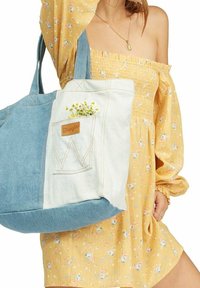 Torba na zakupy z dżinsu z wzorem w kolorze jasnoniebieskim i białym, wyposażona w przednią kieszeń naszytą oraz brązowy skórzany detal z logo. W kieszeni znajdują się małe kwiaty.