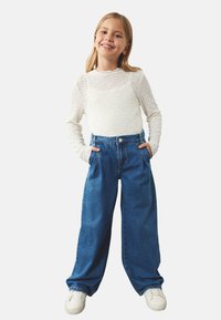 Hvid langærmet bluse med struktureret mønster og rund hals, parret med vide blå denimjeans og hvide sneakers, hænderne i lommerne.