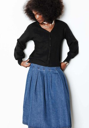 Femme avec une coiffure afro portant un cardigan noir boutonné, des colliers superposés et une jupe midi plissée en denim bleu, posant devant un fond blanc.