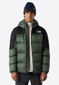 Grön och svart puffjacka med huva, med kviltade sektioner, dragkedja och logotyp på övre vänstra sidan. Bärs över en krämfärgad hoodie.