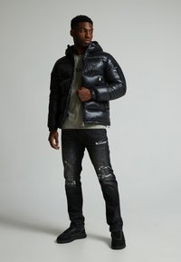 Polstret svart jakke med hette, glidelås foran og elastiske mansjetter, kombinert med revne svarte jeans og svarte sneakers. En ensfarget grønn t-skjorte under.