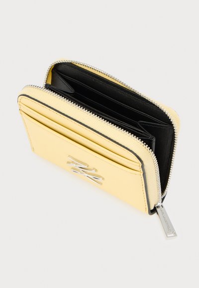 Portefeuille en cuir jaune avec fermeture éclair et éléments en métal argenté, doté d'un intérieur noir, de compartiments pour cartes et d'un logo bien visible sur le devant.