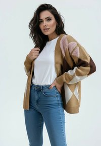 Kobieta ubrana w biały top, niebieskie dżinsy i brązowy kardigan w wzory, pozująca z jedną ręką na biodrze, a drugą trzymającą kardigan.