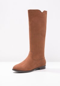 Botte haute jusqu'au genou en daim marron avec un bout arrondi, un talon plat et des détails discrets de couture. Texture lisse avec un design simple et épuré.