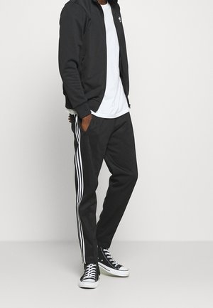 Zwarte jas met ritssluiting en hoge kraag, gecombineerd met zwarte broek met drie witte verticale strepen. Zwarte sneakers maken de outfit compleet.