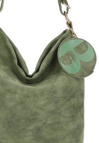 Borsa a spalla in suede verde oliva morbido con un'etichetta verde rotonda attaccata alla tracolla con la scritta "Brigitte" e un gancio in metallo.