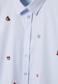 Jasnoniebieska koszula z różnymi haftowanymi wzorami: brązowy niedźwiedź, niebieski kwiat i różowe serce. Posiada klasyczny kołnierzyk i zapięcie na guziki.
