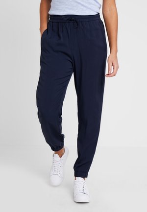 Person walking in navy jogger pants with elastic waistband and cuffs, paired with white sneakers and a tucked-in light gray top.