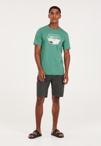 Grøn bomulds t-shirt med et grafisk logo, parret med mørke shorts og sorte slidesandaler. Enkel design, afslappet pasform og komfortabelt udseende.