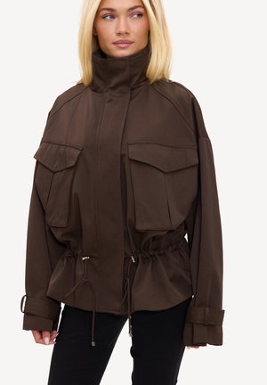 Femme blonde portant une veste marron foncé avec de grandes poches avant, un col haut, une taille à cordon et de longues manches avec des détails de boucle.