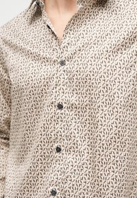 Μπεζ πουκάμισο με κουμπιά, με επαναλαμβανόμενο μοτίβο από μαύρα και καφέ γράμματα "X" και "F", ο γιακάς ανοιχτός, αποκαλύπτοντας τον λαιμό και το ανώτερο μέρος του στήθους.