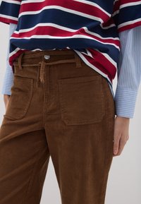 Pantalones de pana marrón con bolsillos frontales y cierre de botón. Llevados con una camisa a rayas roja, blanca y azul marino y una camisa de rayas azules debajo.