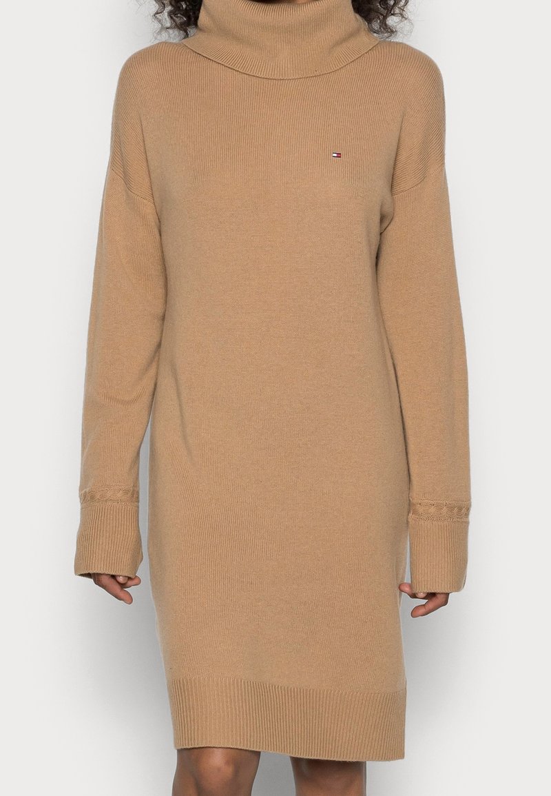 Personne portant une robe-pull à col roulé beige à manches longues avec un petit logo sur la poitrine et des poignets et un ourlet côtelés.