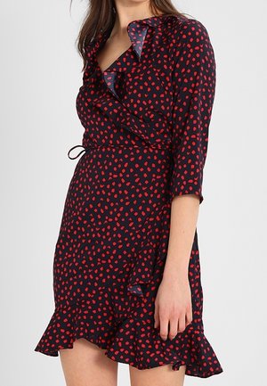 Woman wearing a black wrap dress with red irregular polka dots, ruffled hem, and three-quarter sleeves, standing against a plain background.