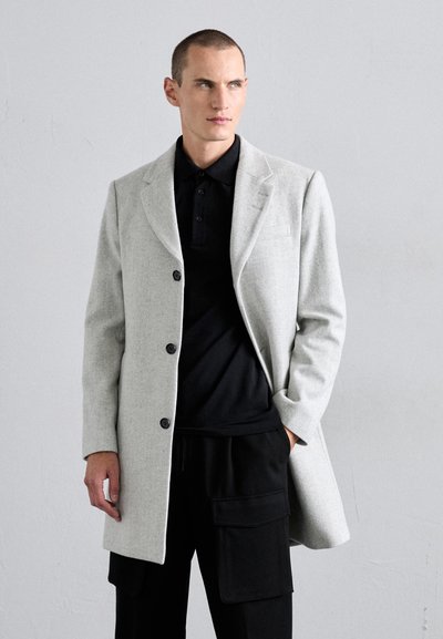Light gray wool overcoat with a structured design, single-breasted front, and black buttons, worn over a black polo shirt and black pants.