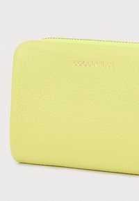 Vaaleankeltainen pinnattu nahkainen lompakko, jossa on kultainen vetoketju ja kohokuvioitu kultainen brändinimi "COCCINELLE" edessä.