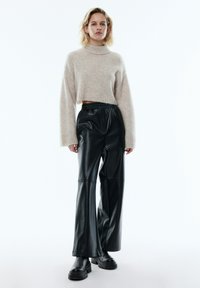 Hellbeige abgeschnittenen Pullover mit einem hohen Kragen und weiten Ärmeln, kombiniert mit schwarzen, weiten Kunstlederhosen und klobigen schwarzen Stiefeln.