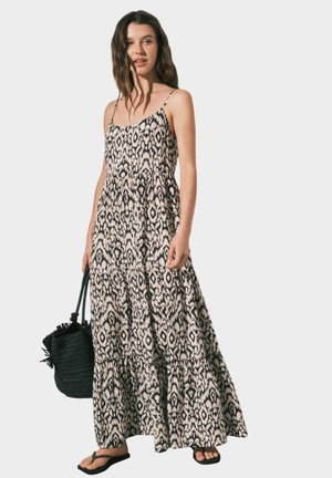 Giovane donna con abito lungo senza maniche a fantasia bianco e nero, che tiene una borsa nera intrecciata, indossa sandali neri su uno sfondo semplice.