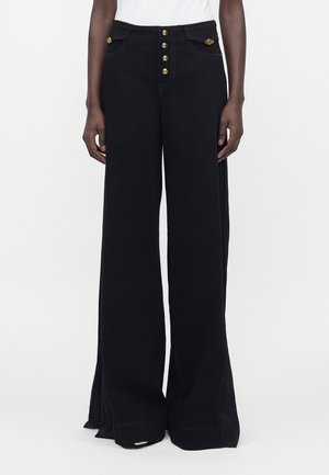 Osoba nosí čierne široké nohavice s vysokým pásom, so zlatým zapínaním na gombíky a dekoratívnymi zlatými detailmi na predných vreckách.