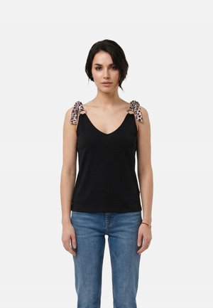 Donna che indossa una canotta nera senza maniche con spalline a fiocco stampa leopardata e jeans blu, in piedi contro uno sfondo chiaro e semplice.