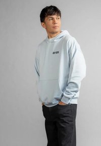 Młody mężczyzna ubrany w jasnoniebieską bluzę z kapturem z napisem "WILLIAMS EST. 1977", stoi z rękami w kieszeniach, patrząc lekko na bok.