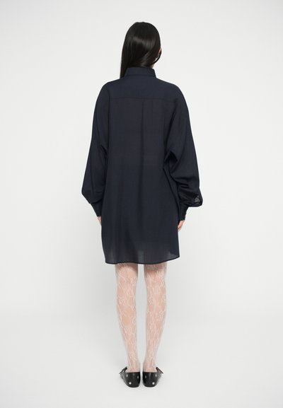 Vestido camisa oversized azul-marinho com mangas longas, colarinho abotoado e tecido translúcido, usado sobre meias optimizadas com padrão de renda claras e sapatos pretos.