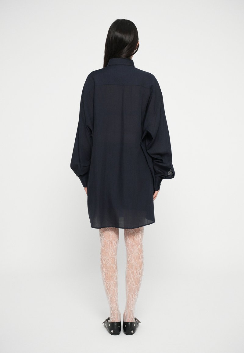 Navyblaues Oversized-Hemdkleid mit langen Ärmeln, Knopfleiste und transparenter Stoff, getragen über hellen, spitzenbesetzten Strumpfhosen und schwarzen Schuhen.