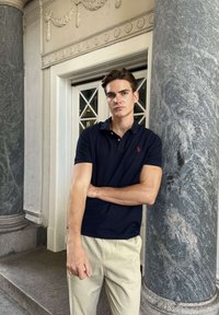 Tânăr cu piele deschisă la culoare și păr castaniu, sprijinit de un stâlp de marmură, purtând un tricou polo bleumarin și pantaloni bej lângă intrarea ornamentată a unei clădiri.