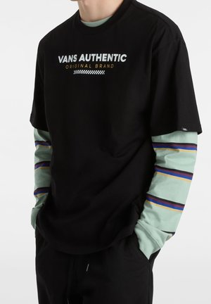 Person trägt ein schwarzes Vans Authentic T-Shirt über einem grünen Langarmshirt mit blauen, gelben und violetten Streifen, Hände in den Taschen.