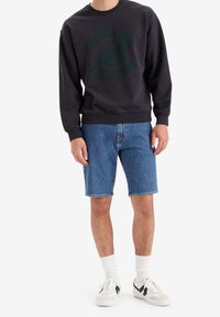 Man som bär en svart sweatshirt med texten "We Are All Star Dust" och en planetgrafik, blå denimshorts, vita strumpor och vita sneakers.