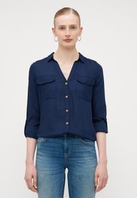 Chemise boutonnée bleu marine avec un col en V, deux poches poitrine et des manches retroussées. Tissu lisse et boutons dorés ajoutent une touche de détail. Associée à un jean bleu.