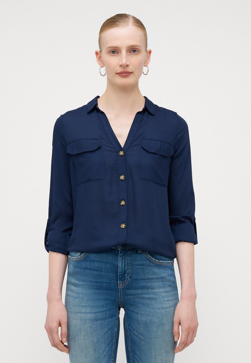 Chemise boutonnée bleu marine avec un col en V, deux poches poitrine et des manches retroussées. Tissu lisse et boutons dorés ajoutent une touche de détail. Associée à un jean bleu.
