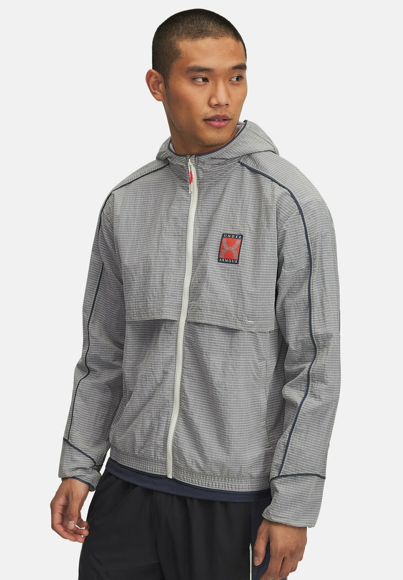 Graue leichte Jacke mit Kapuze, ausgestattet mit einem Frontreißverschluss, kontrastierenden marineblauen Besätzen und einem Logo auf der Brust. Strukturierter Stoff mit kariertem Muster.