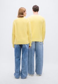 Κίτρινα πλεκτά πουλόβερ με ριμπ μανσέτες και τελείωμα, συνδυασμένα με φαρδιά μπλε τζιν παντελόνια, εμφανιζόμενα από πίσω με φόντο λευκό.